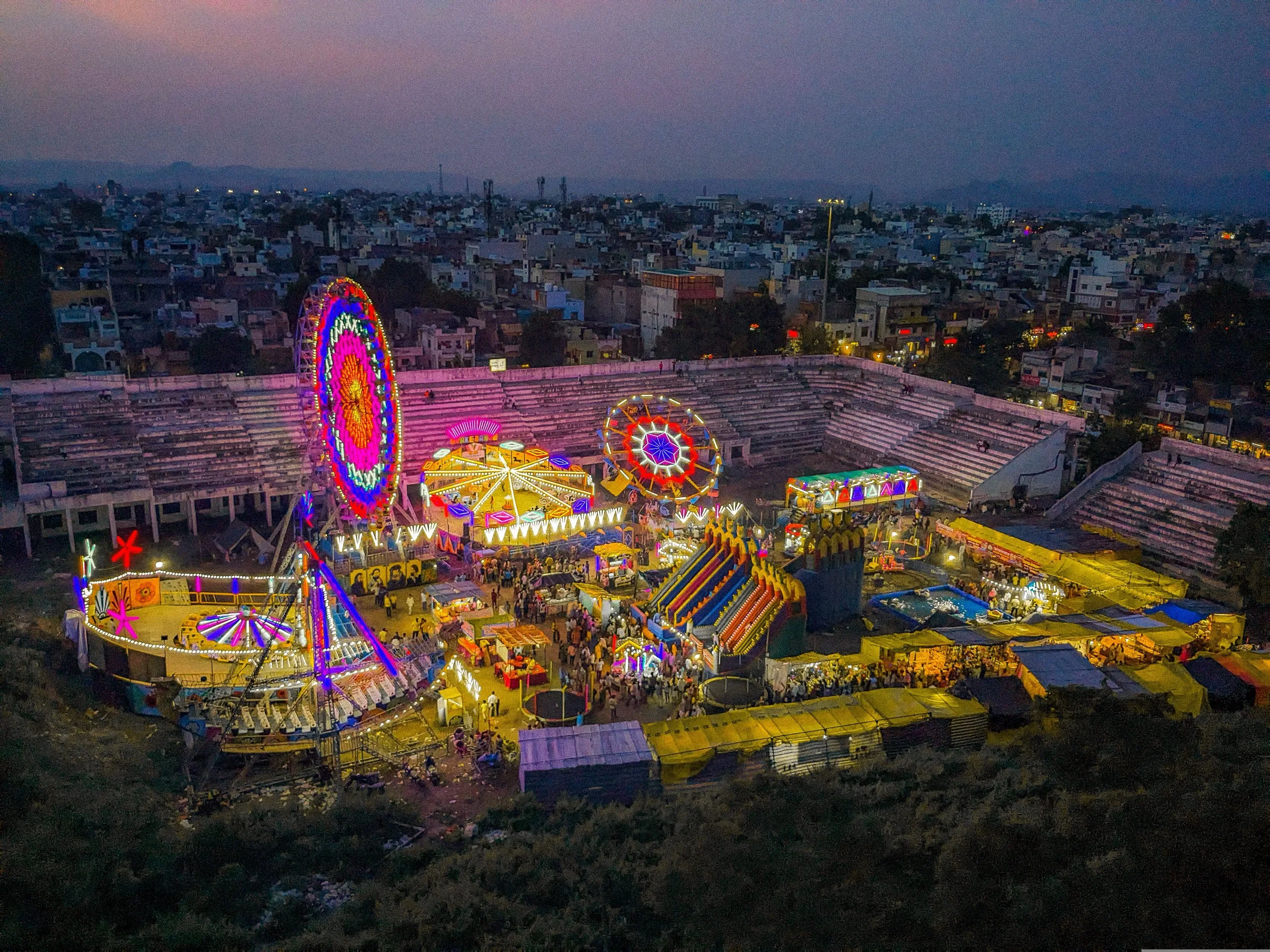 A fairground lit up at dusk, with Ferris wheels, rides and stalls