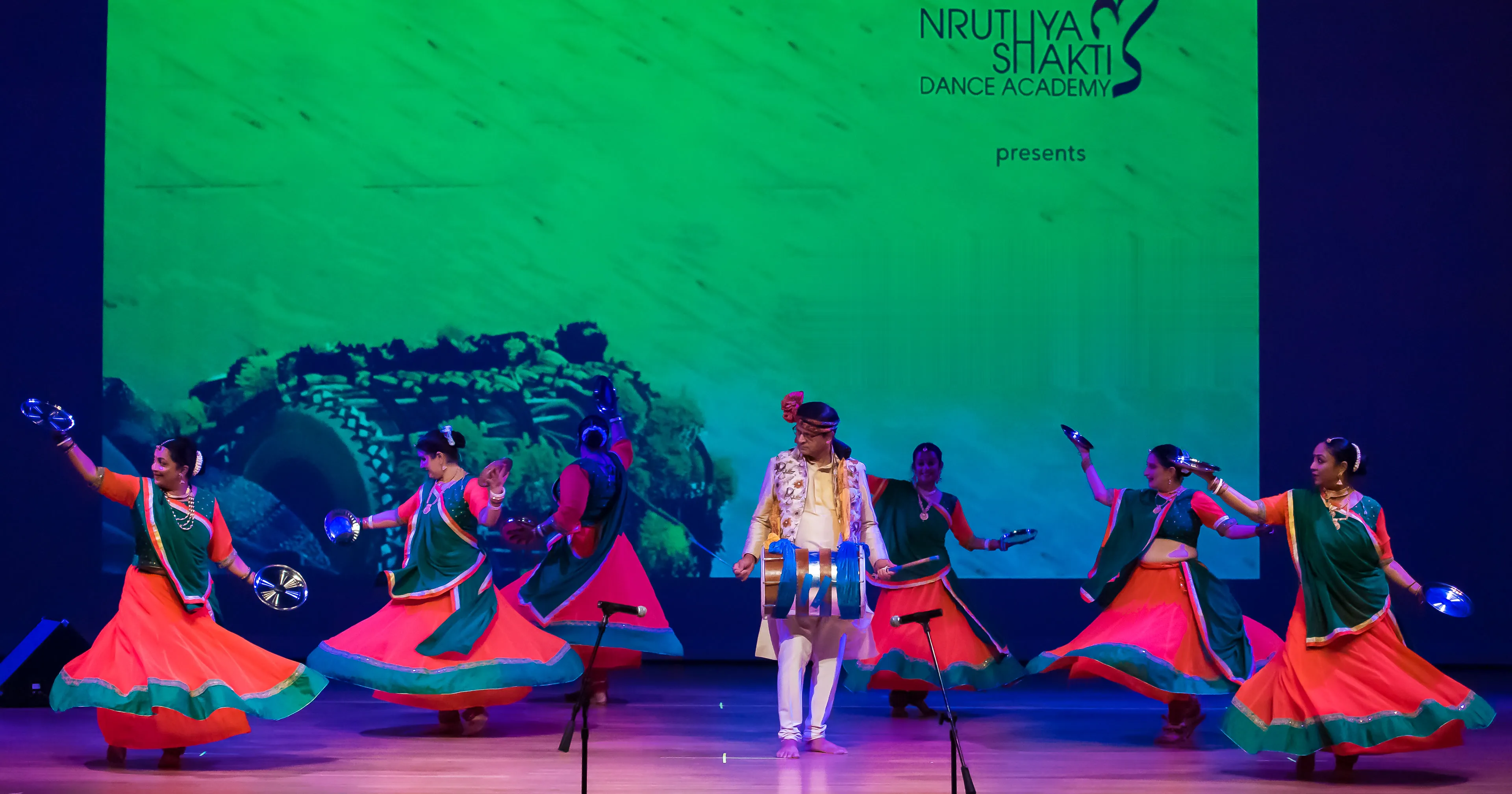 NSDA dancers in orange and green, cymbals in hand, dancing Hudo around a drummer at Loknritya Dhwani