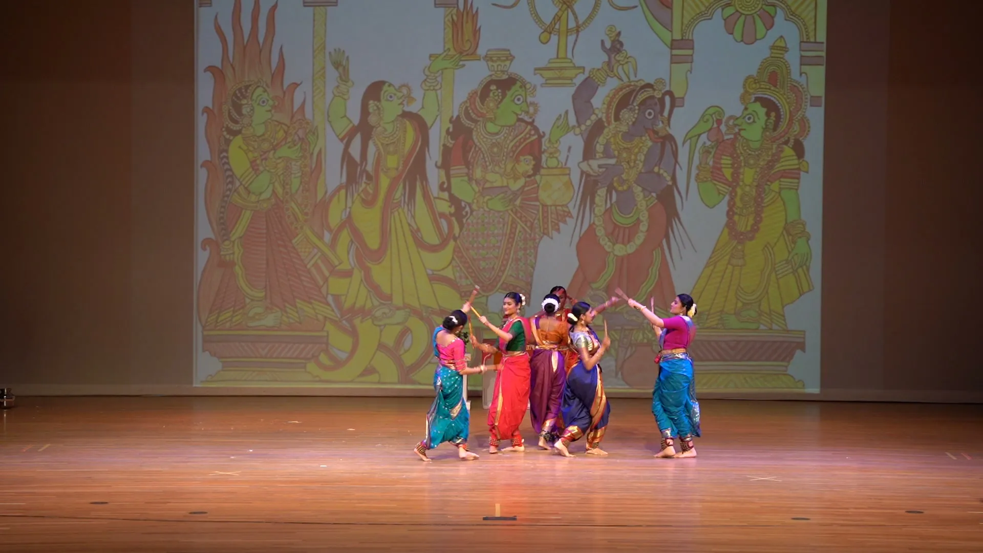 NSDA dancers in sarees on stage, sticks raised overhead, before a painted backdrop