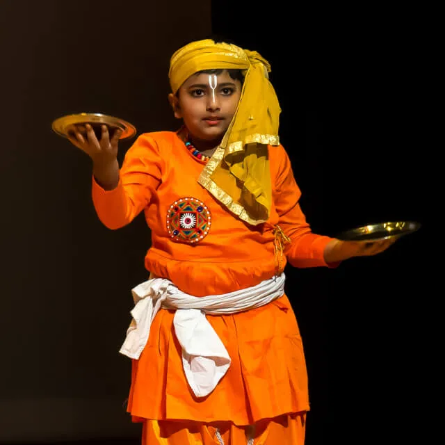 A junior Elite dancer in saffron, holding two brass plates